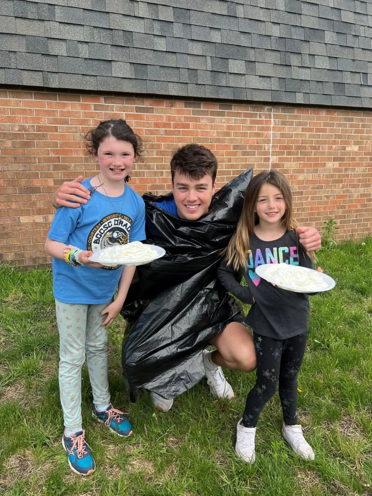 Two girls holding plates of whipped cream covered pies and a BGCStory staff member in a larg black trashbag.