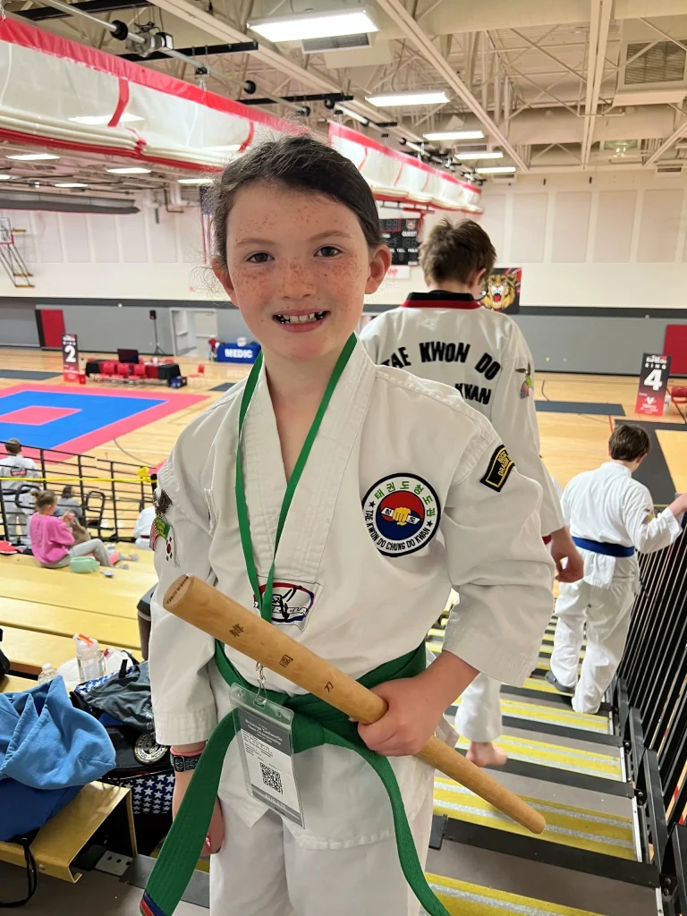 A young girl in Taekwondo outfit standing on bleachers in a gym.